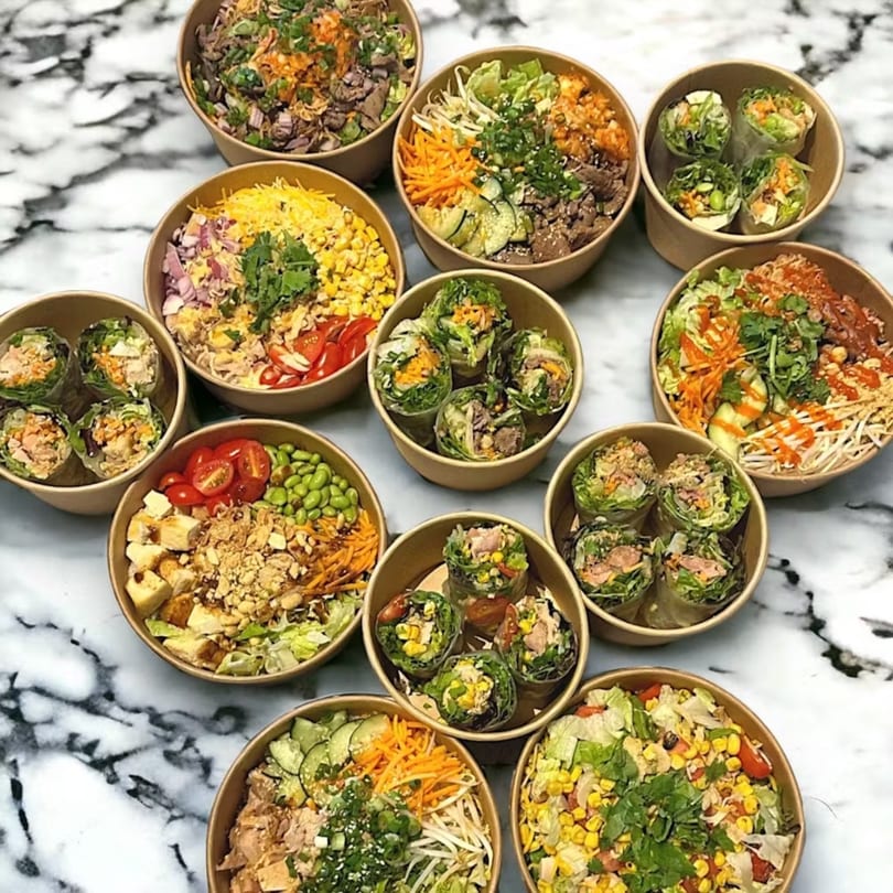 An overhead view of multiple Asian-inspired bowls and spring rolls on a marble surface.