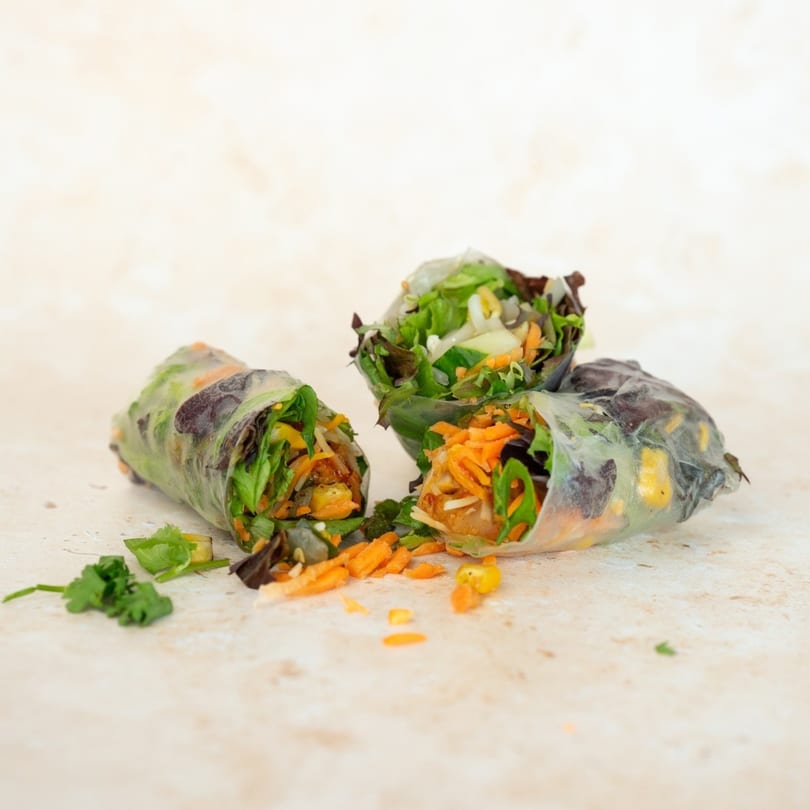 Three fresh Up Leaf Café spring rolls, filled with vibrant vegetables, on a light-colored surface.