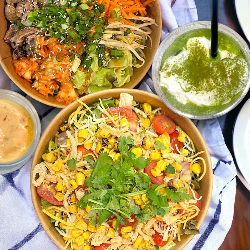 Two vibrant Asian-inspired bowls, one with beef and kimchi, the other with corn and tomatoes, alongside a green drink.